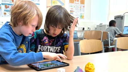 Two children using a tablet at a classroom table, with 3D‑printed models nearby and other students working at computers in the background.