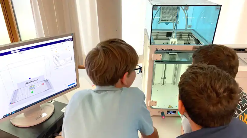 Children watching a 3D printer while a 3D model is prepared on a nearby computer screen.