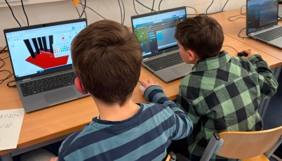Two students at laptops designing and coding, with a 3D model on one screen and block-based code on another.