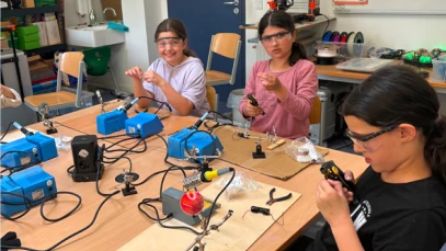 Students soldering electronics at a workshop table with soldering stations and tools.